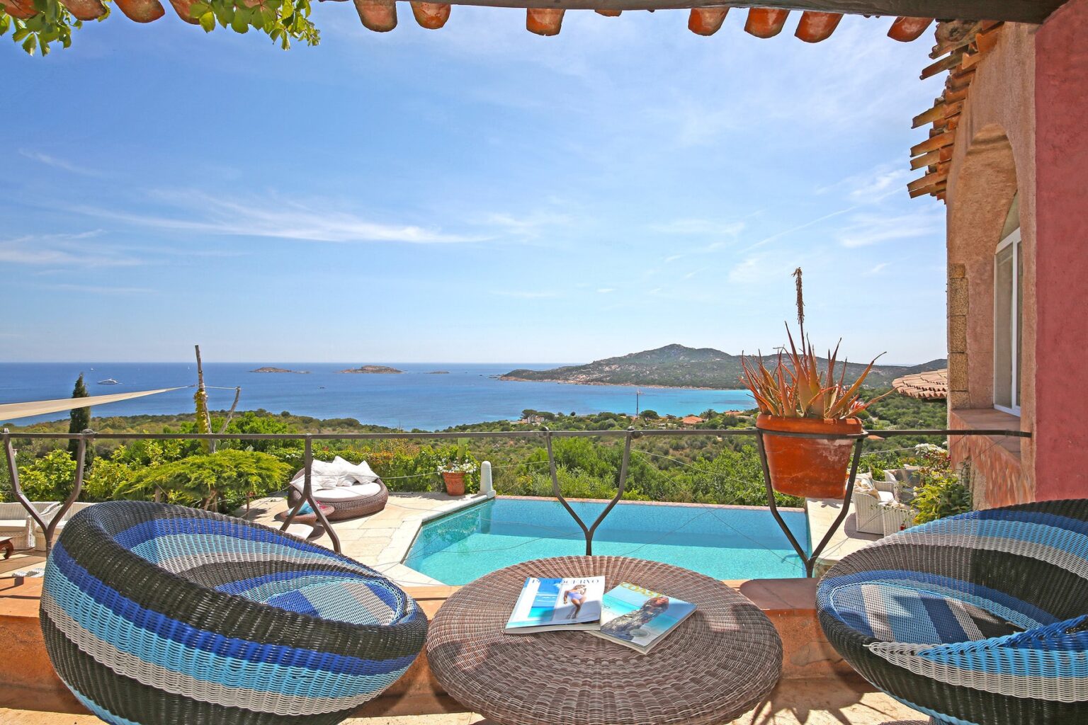 Scenic view from a terrace featuring two colorful chairs, a round table with magazines, and a swimming pool overlooking a beautiful coastline with islands in the distance and clear blue skies. Ideal for vacation rentals and luxury getaways. | Only on SopranoVillas