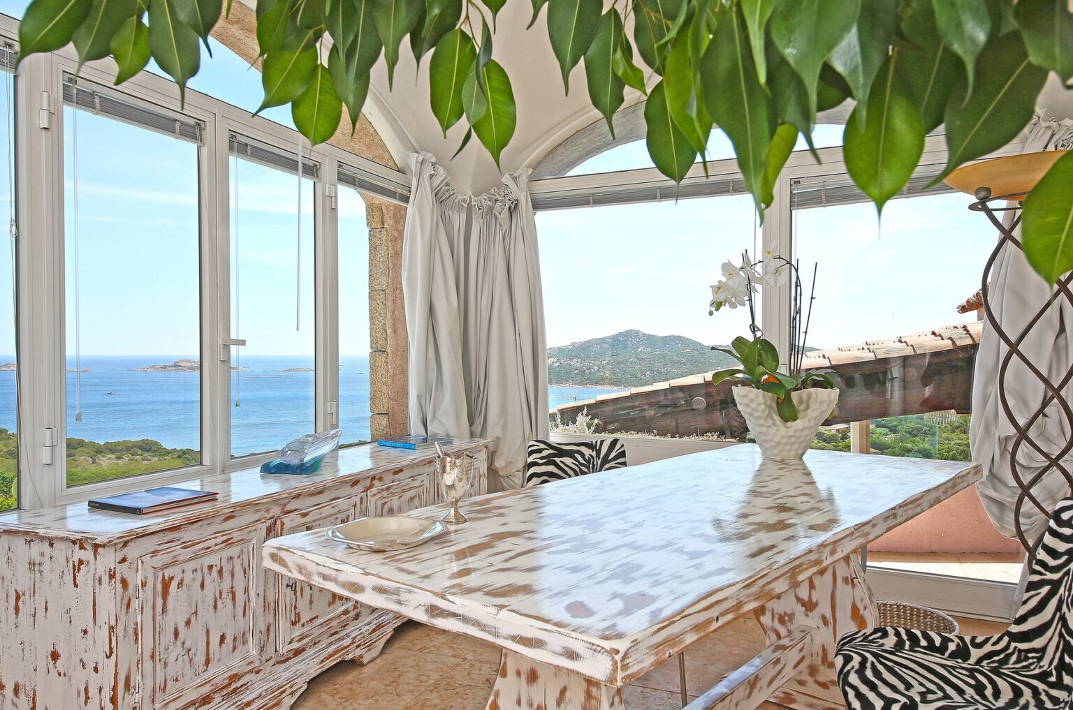 Bright dining area with large windows offering a stunning view of the sea and surrounding hills, featuring a stylish white table, decorative plants, and elegant decor. | Only on SopranoVillas