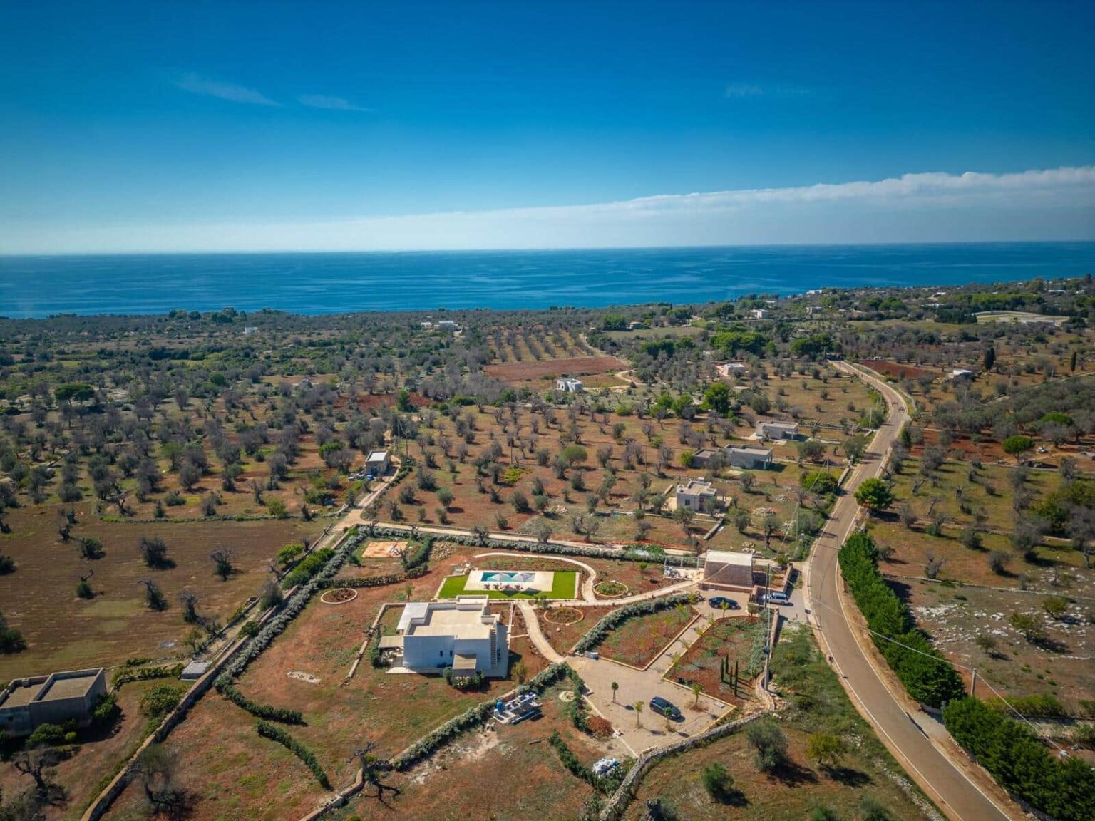 Aerial view of a coastal landscape featuring olive groves, a modern villa with a pool, and winding roads leading to the sea under a clear blue sky. | Only on SopranoVillas