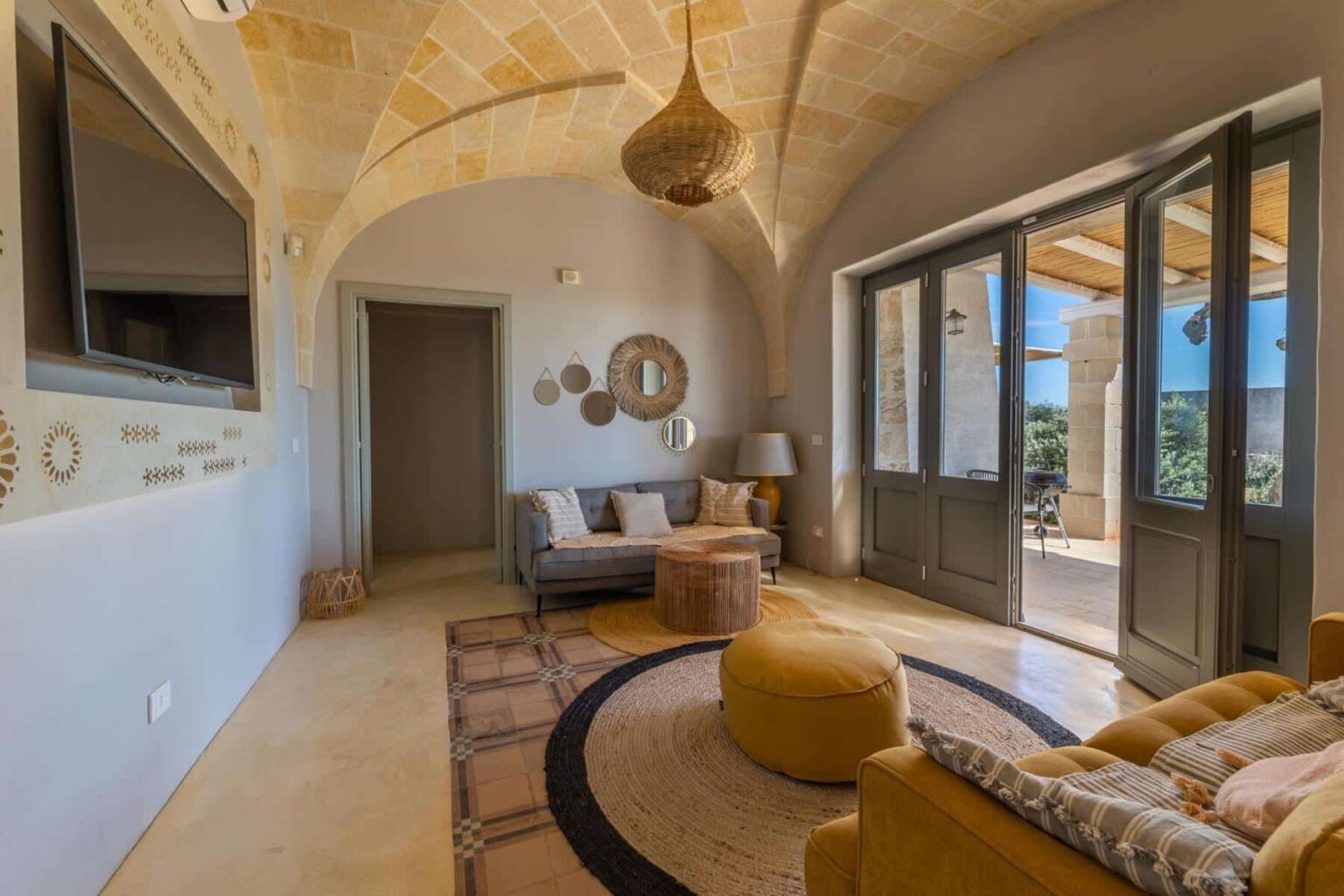 Cozy living room with arched stone ceiling, featuring a grey sofa, yellow pouf, and wooden coffee table. The room includes decorative mirrors, a modern TV, and large glass doors leading to an outdoor patio with a scenic view. Natural light enhances the warm, inviting atmosphere. | Only on SopranoVillas