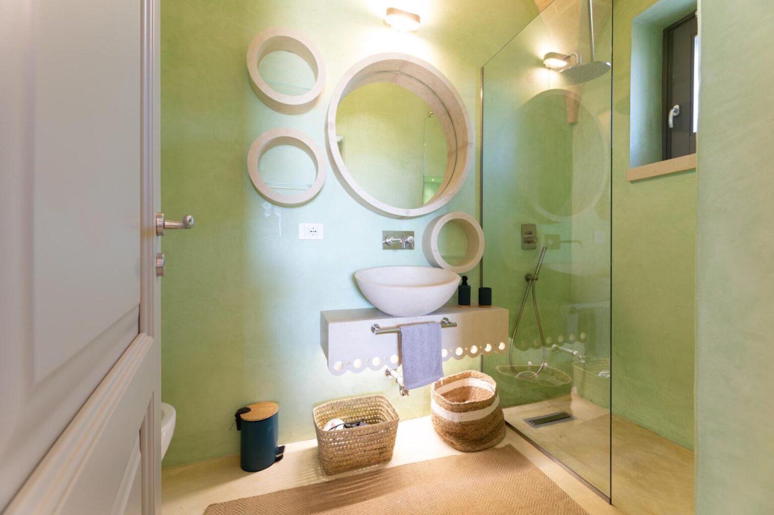 Modern bathroom interior featuring a green textured wall, a round mirror, a stylish sink, and a glass shower. Decorative baskets and a soft rug add warmth to the space. | Only on SopranoVillas