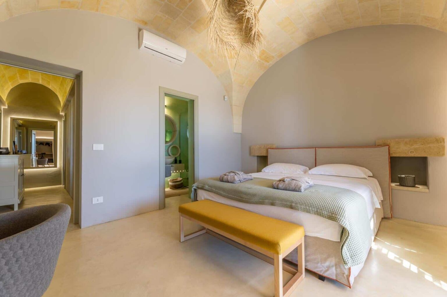 Modern bedroom interior featuring a double bed with white linens, a textured green throw, and decorative towels. The room has a warm, arched ceiling, stylish yellow bench, and a cozy armchair. A doorway leads to a bathroom area, enhancing the spacious and inviting atmosphere. Ideal for relaxation and comfort. | Only on SopranoVillas