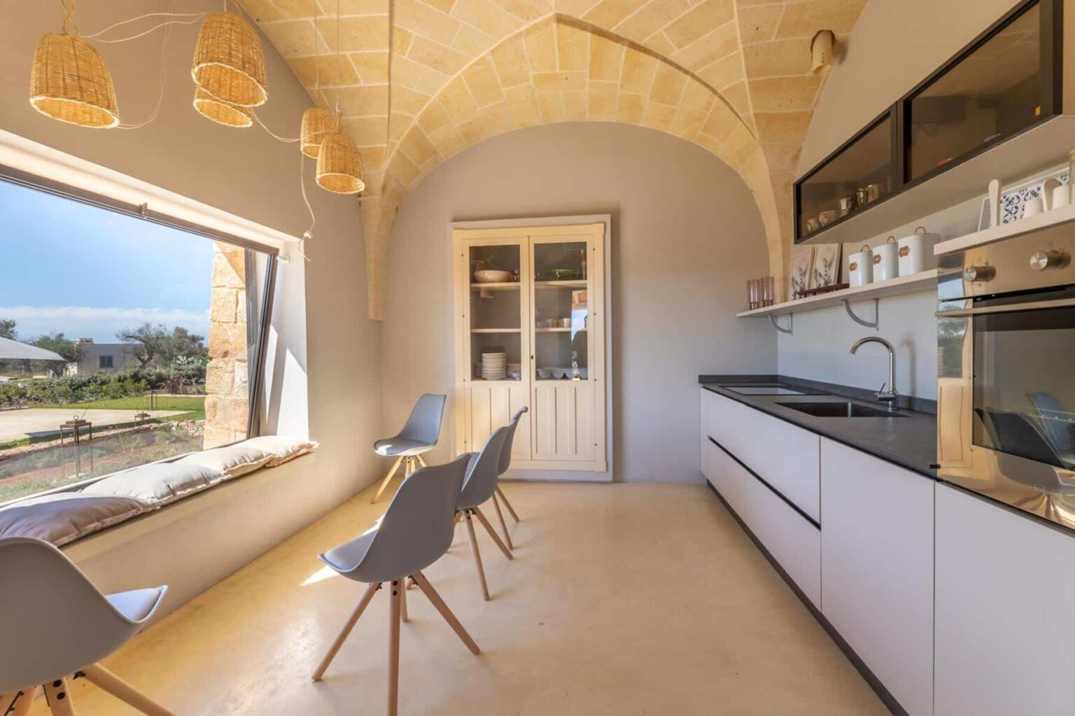 Modern kitchen interior featuring minimalist design, stone archway, sleek cabinetry, and large window with scenic views. The space includes stylish chairs and contemporary appliances, creating a bright and inviting atmosphere. | Only on SopranoVillas