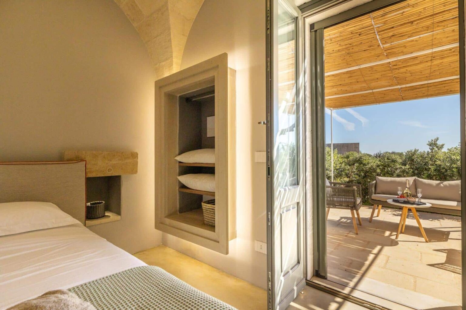 Modern bedroom with a cozy atmosphere, featuring a built-in storage space for pillows and an open door leading to a sunlit terrace with outdoor seating. Ideal for relaxation and enjoying scenic views. | Only on SopranoVillas