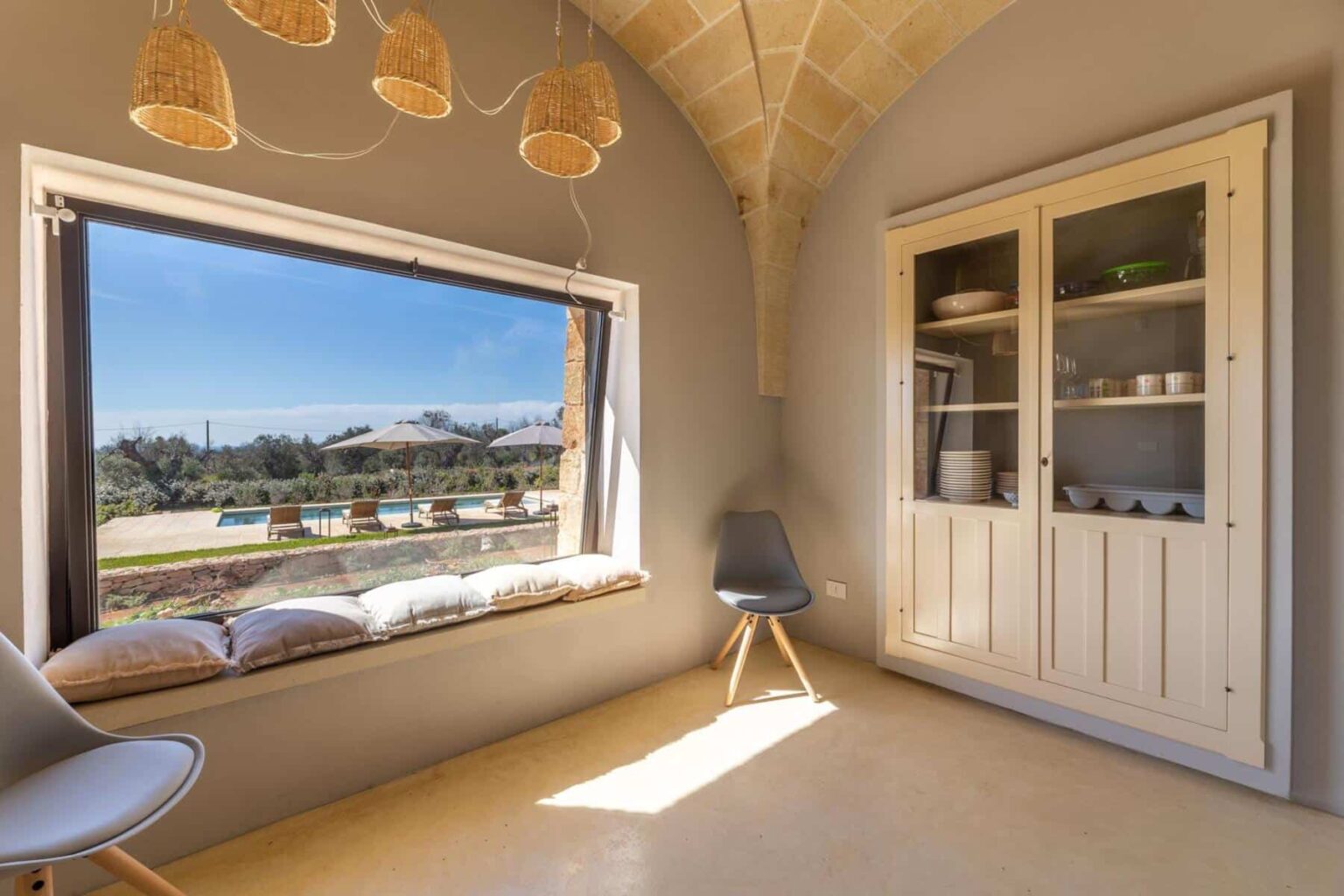 Cozy interior space featuring a large window with a scenic view, a comfortable window seat with cushions, a modern chair, and a stylish cabinet displaying dishware. Natural light fills the room, highlighting the architectural details and warm color palette. | Only on SopranoVillas