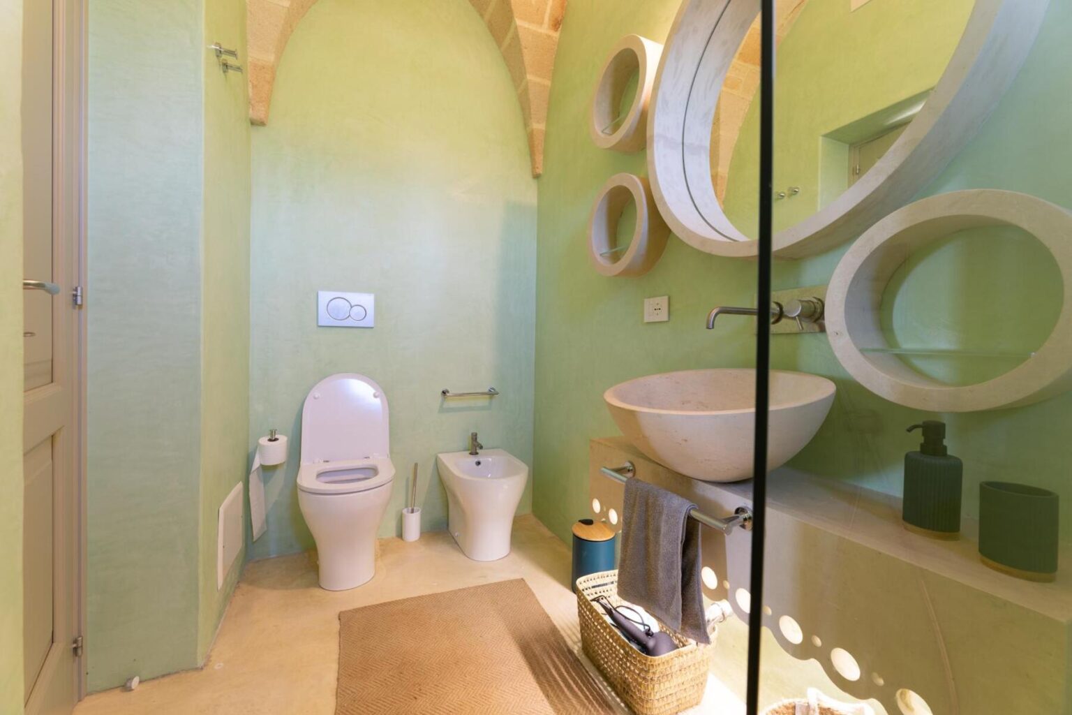 Modern bathroom featuring a green color scheme, a wall-mounted toilet, bidet, and a stylish round basin. The space is illuminated with natural light, highlighting circular mirrors and minimalistic decor. | Only on SopranoVillas