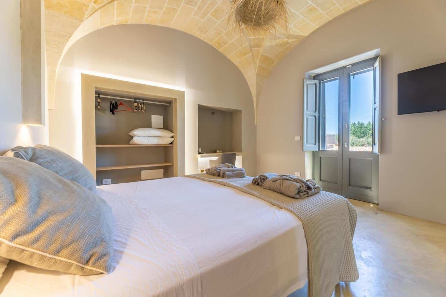 Modern bedroom with an arched ceiling, featuring a comfortable bed, stylish linens, and natural light streaming through large windows. The room includes a minimalist closet and a flat-screen TV, creating a cozy and inviting atmosphere. | Only on SopranoVillas