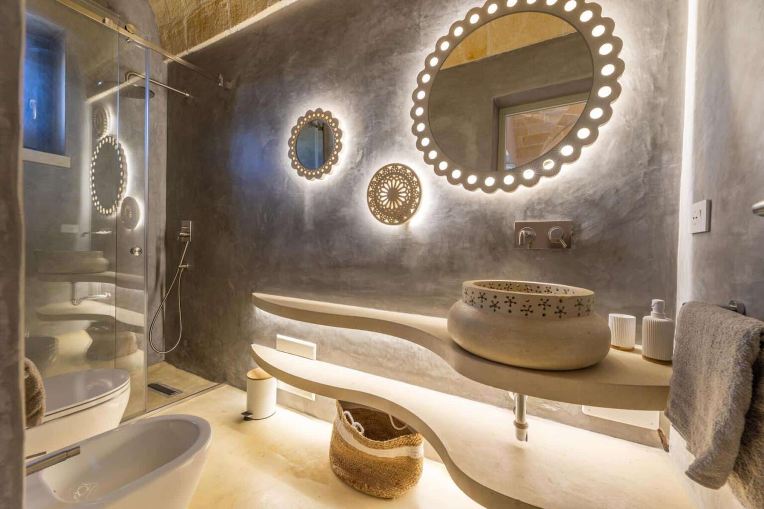 Modern bathroom interior featuring sleek design elements, including a glass shower, decorative mirrors with LED lighting, a stylish sink on a curvy countertop, and neutral color tones for a contemporary aesthetic. | Only on SopranoVillas