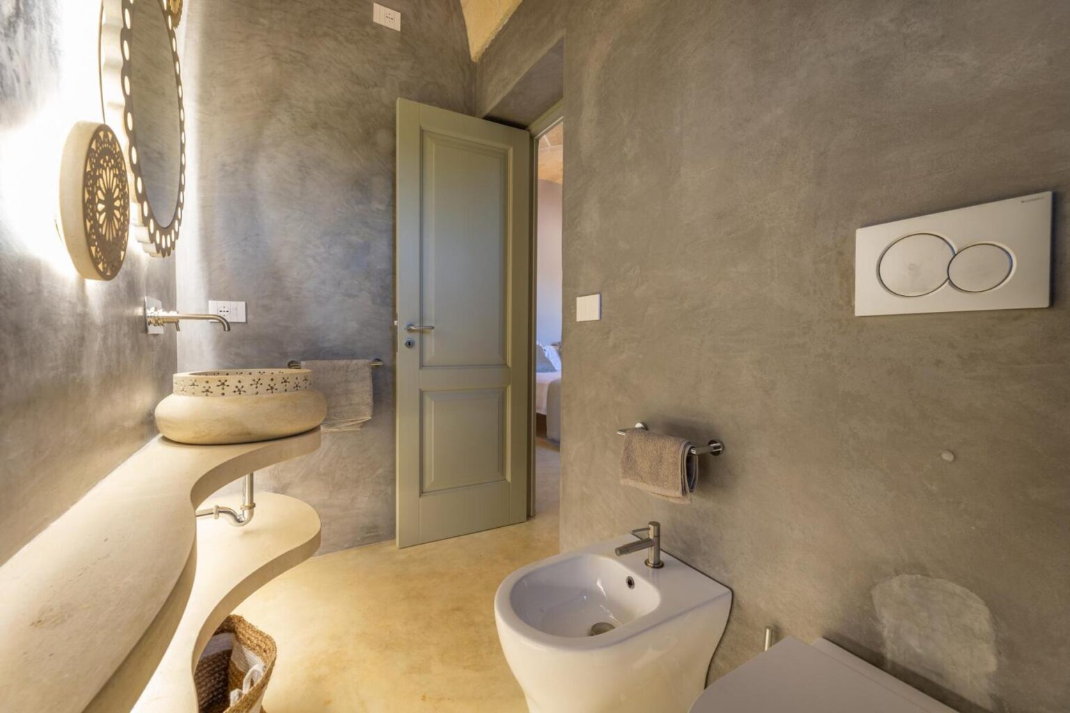 Modern bathroom interior featuring a sleek design with a gray wall finish, a contemporary toilet, and a stylish washbasin. A wooden door leads to another room, and decorative elements enhance the aesthetic. Ideal for showcasing minimalist bathroom decor ideas. | Only on SopranoVillas