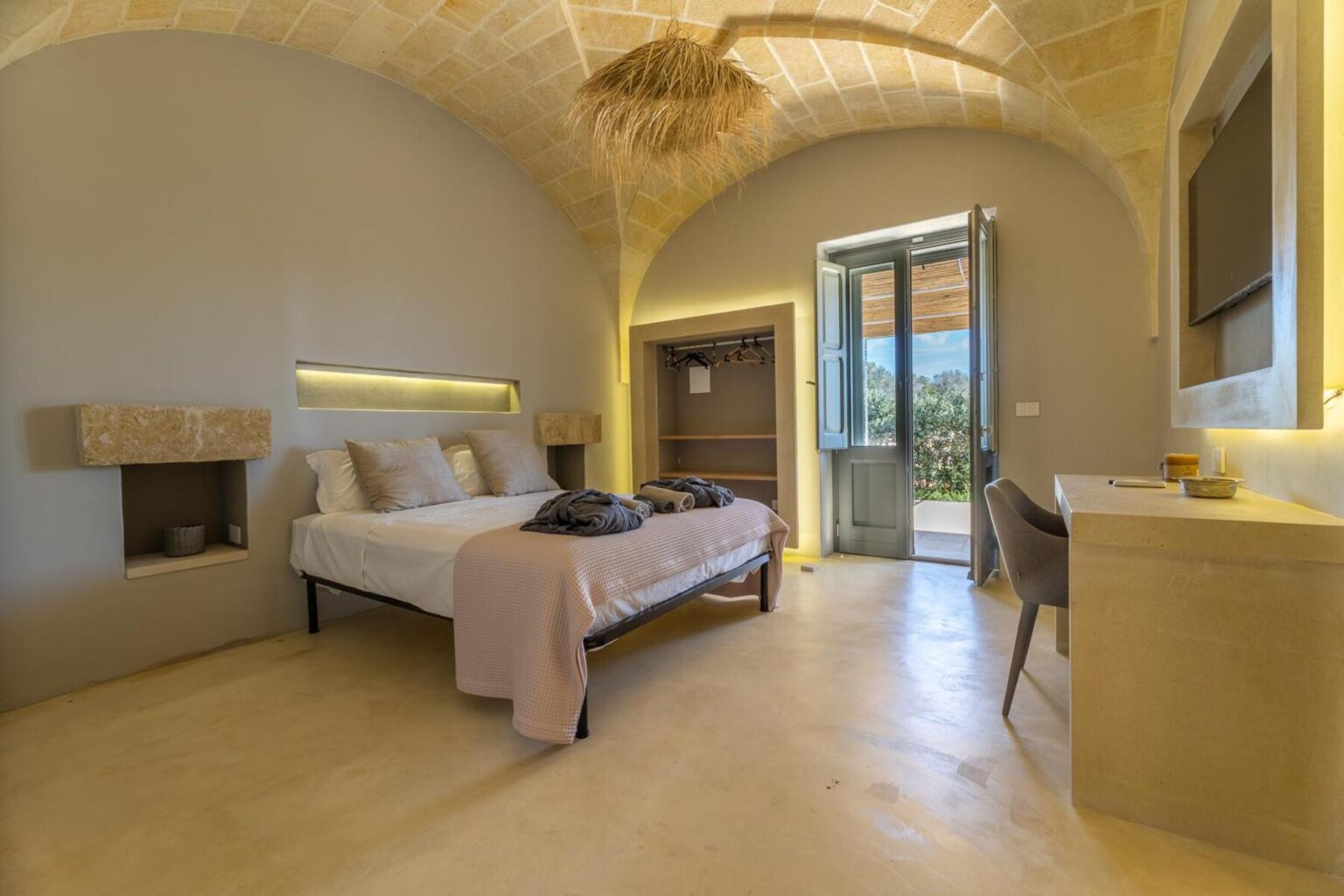 Modern bedroom interior featuring a double bed with neutral linens, minimalist decor, and natural light from an open door leading to a scenic view. The room includes stylish furnishings, an artistic ceiling, and a warm color palette, ideal for a relaxing getaway. | Only on SopranoVillas