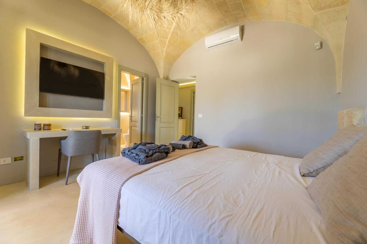 Modern bedroom interior featuring a cozy bed with soft linens, a large flat-screen TV mounted on the wall, and a stylish desk with a chair. The room showcases elegant arches and warm lighting, creating a relaxing atmosphere. | Only on SopranoVillas
