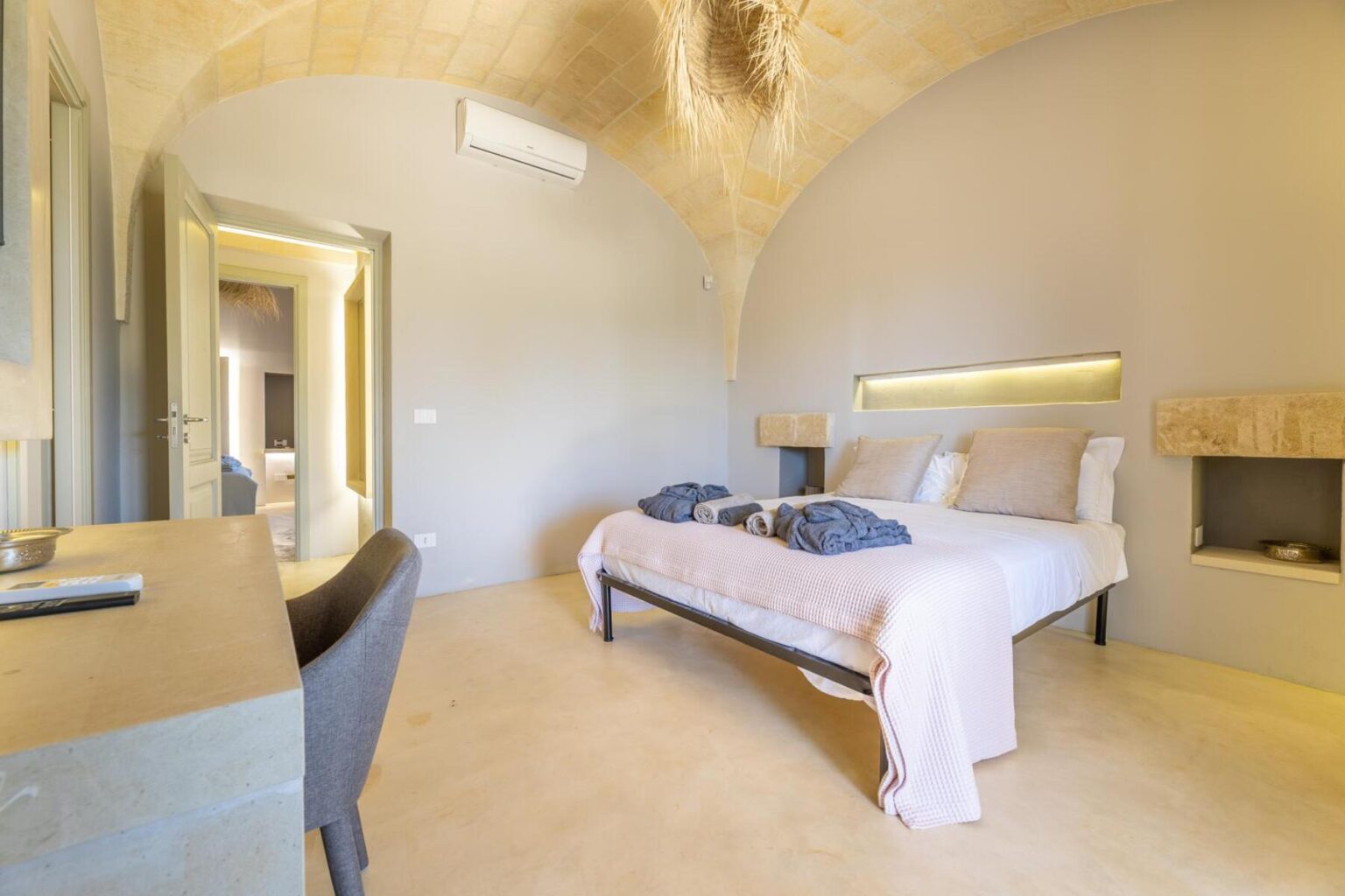 Modern bedroom with an arched ceiling, featuring a double bed with gray towels, minimalist decor, and a stylish desk. The room is well-lit with natural light and has an air conditioning unit for comfort. | Only on SopranoVillas