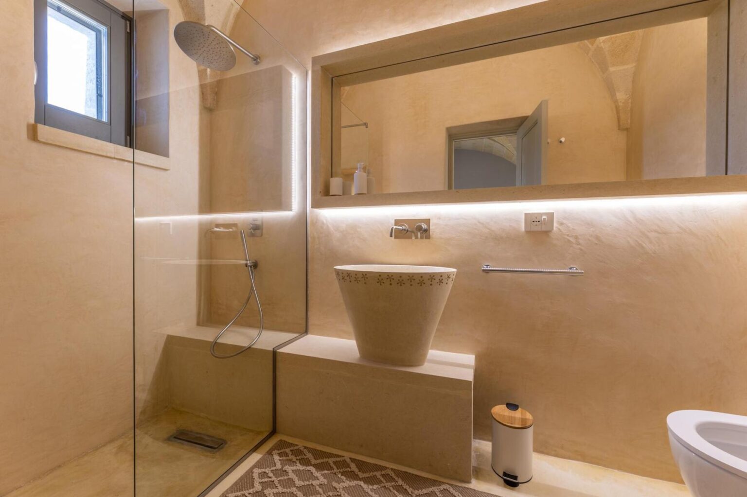 Modern bathroom design featuring a spacious shower area with glass enclosure, a minimalist stone sink, and ambient lighting. The decor includes neutral tones and a stylish rug, creating a serene atmosphere. | Only on SopranoVillas