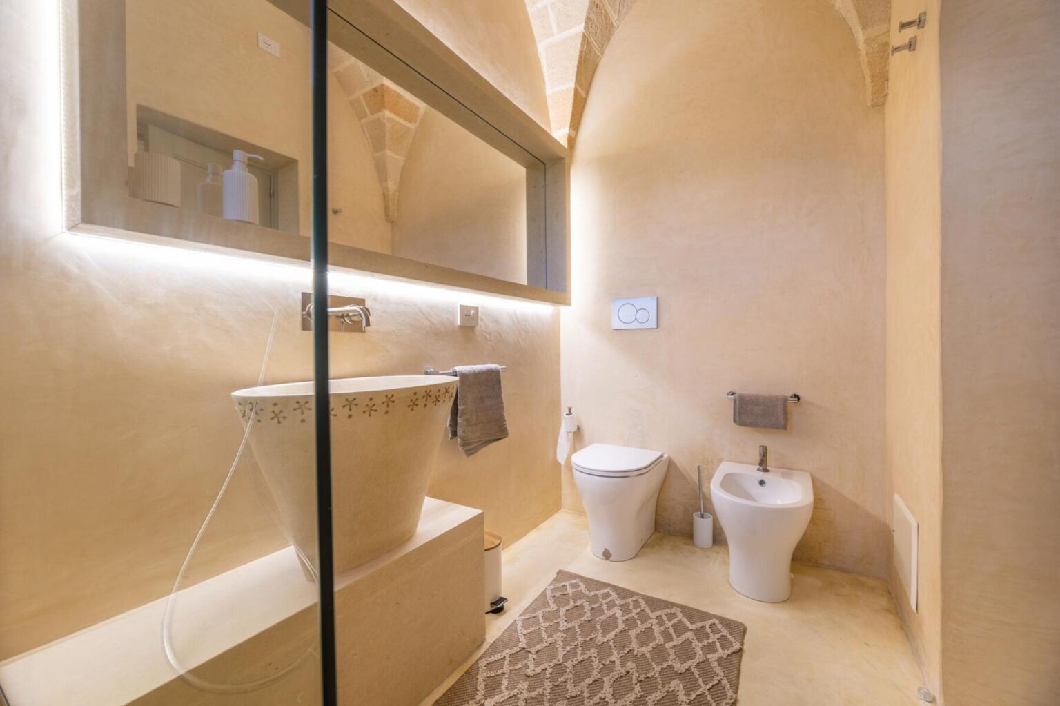 Modern bathroom design featuring a minimalist aesthetic with a stone toilet, bidet, and a large mirror illuminated by soft lighting. The walls are finished in warm beige tones, complemented by a textured area rug. | Only on SopranoVillas