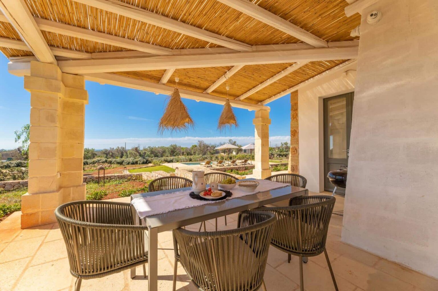 Outdoor dining area with a modern table and chairs under a thatched roof, surrounded by lush greenery and a clear blue sky, showcasing a tranquil setting perfect for relaxation and meals al fresco. | Only on SopranoVillas