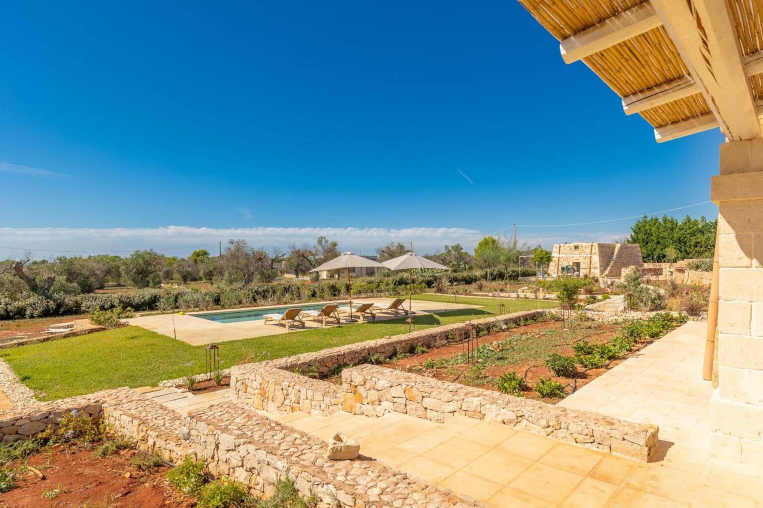 Scenic view of a luxurious villa with a swimming pool, sun loungers, and lush gardens, set against a clear blue sky. The image showcases natural stone landscaping and a serene outdoor living space, ideal for relaxation and leisure. | Only on SopranoVillas