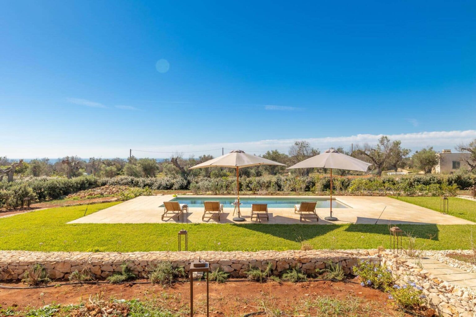 A serene outdoor pool area featuring lounge chairs and umbrellas, surrounded by lush green grass and olive trees under a clear blue sky, ideal for relaxation and leisure activities. | Only on SopranoVillas