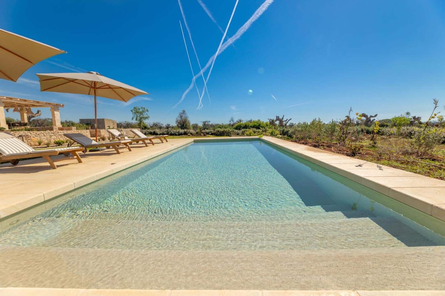Serene outdoor swimming pool with clear water, surrounded by lounge chairs and umbrellas, set against a bright blue sky with contrails, showcasing a tranquil landscape and lush greenery. | Only on SopranoVillas