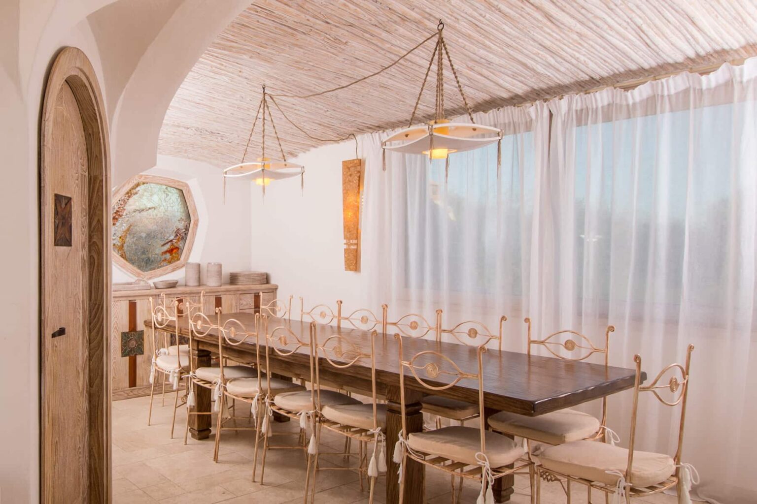 Modern dining room featuring a long wooden table surrounded by elegant chairs, bright natural light streaming through sheer curtains, and stylish pendant lighting. The decor includes a decorative mirror and a minimalist aesthetic, creating a warm and inviting atmosphere. | Only on SopranoVillas
