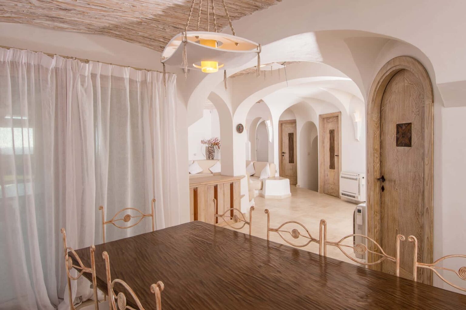 Spacious interior of a modern dining area featuring a wooden table with ornate chairs, sheer white curtains, and arched doorways leading to a cozy living space. Natural light illuminates the room, enhancing the minimalist design and warm wooden accents. | Only on SopranoVillas