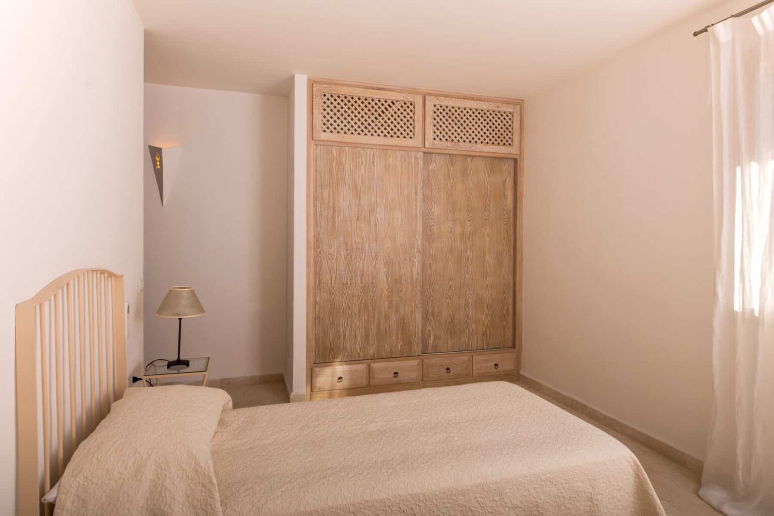 Cozy bedroom featuring a light-colored bedspread, wooden headboard, and a spacious wardrobe with a natural finish. Soft lighting from a bedside lamp enhances the serene atmosphere, complemented by neutral wall colors and a sheer curtain allowing natural light. Ideal for relaxation and comfort. | Only on SopranoVillas