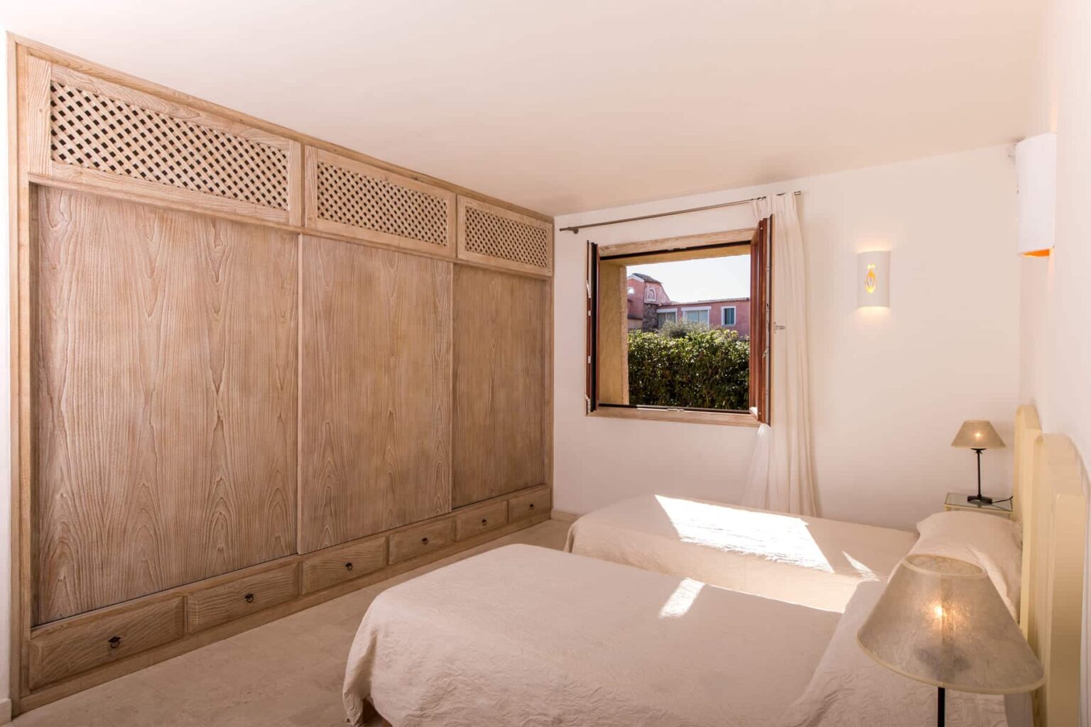 Cozy bedroom featuring two single beds, a wooden wardrobe with intricate lattice design, and a window allowing natural light to illuminate the space. The decor is minimalistic and inviting, with a bedside lamp adding a warm touch. | Only on SopranoVillas
