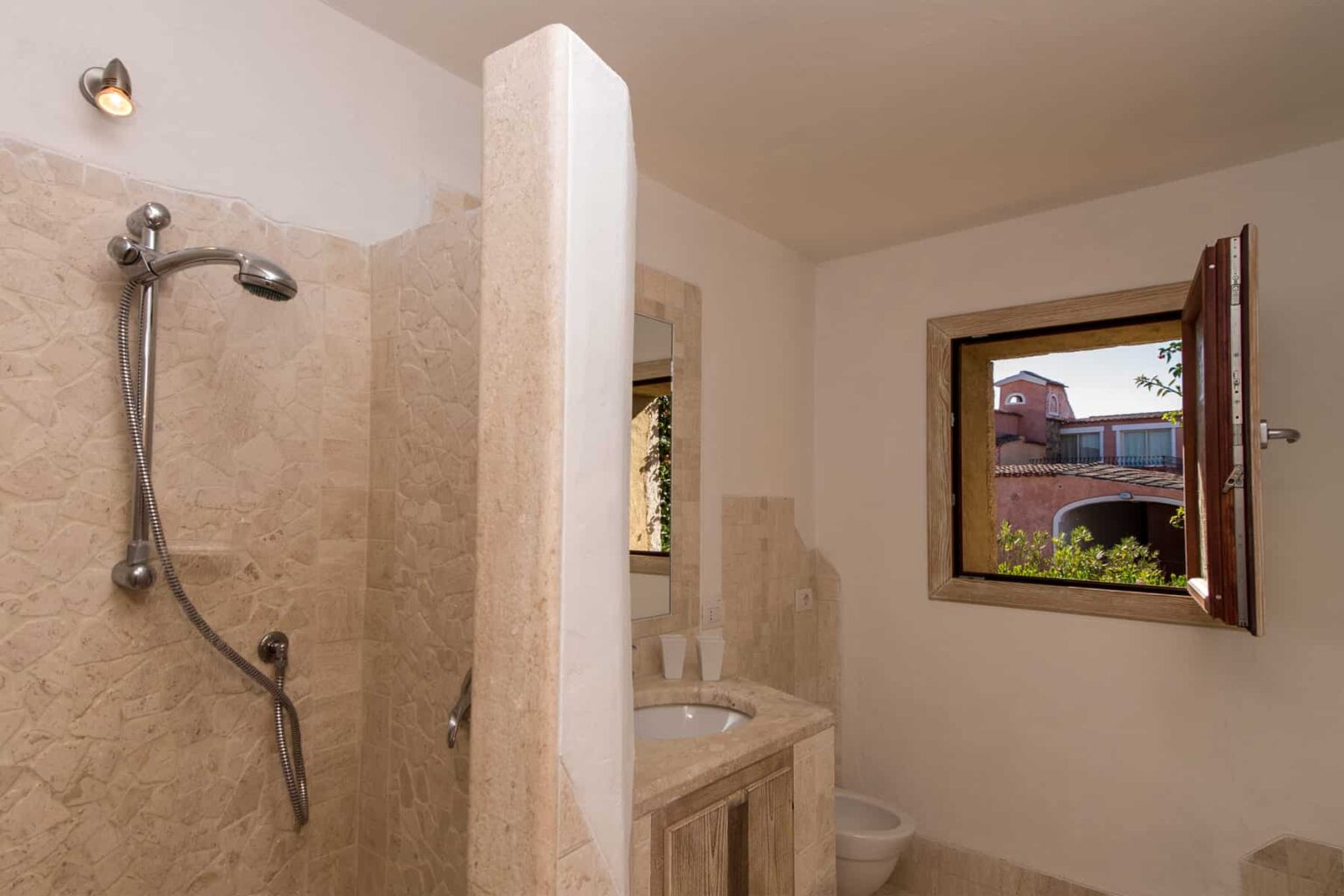 Modern bathroom featuring a spacious shower area with a sleek showerhead, beige stone walls, and a wooden window overlooking a garden. The room is well-lit and designed for comfort and relaxation. | Only on SopranoVillas
