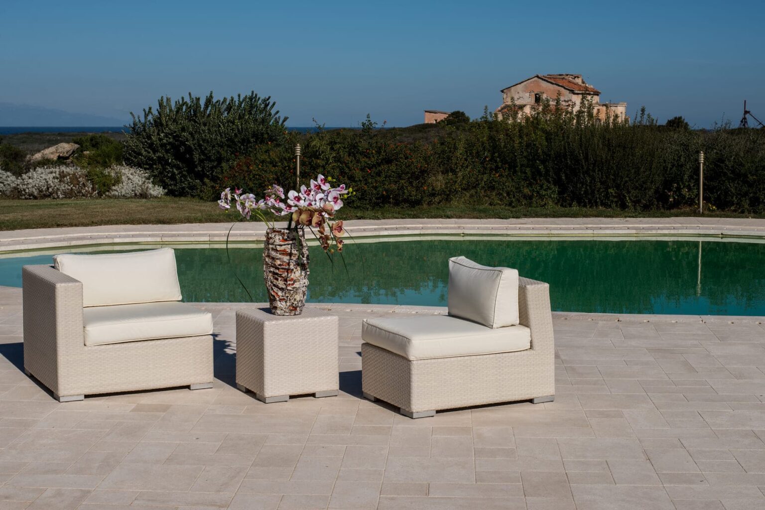 Modern outdoor seating area with two white chairs and a side table, featuring a vase of flowers, beside a tranquil pool and scenic landscape. | Only on SopranoVillas