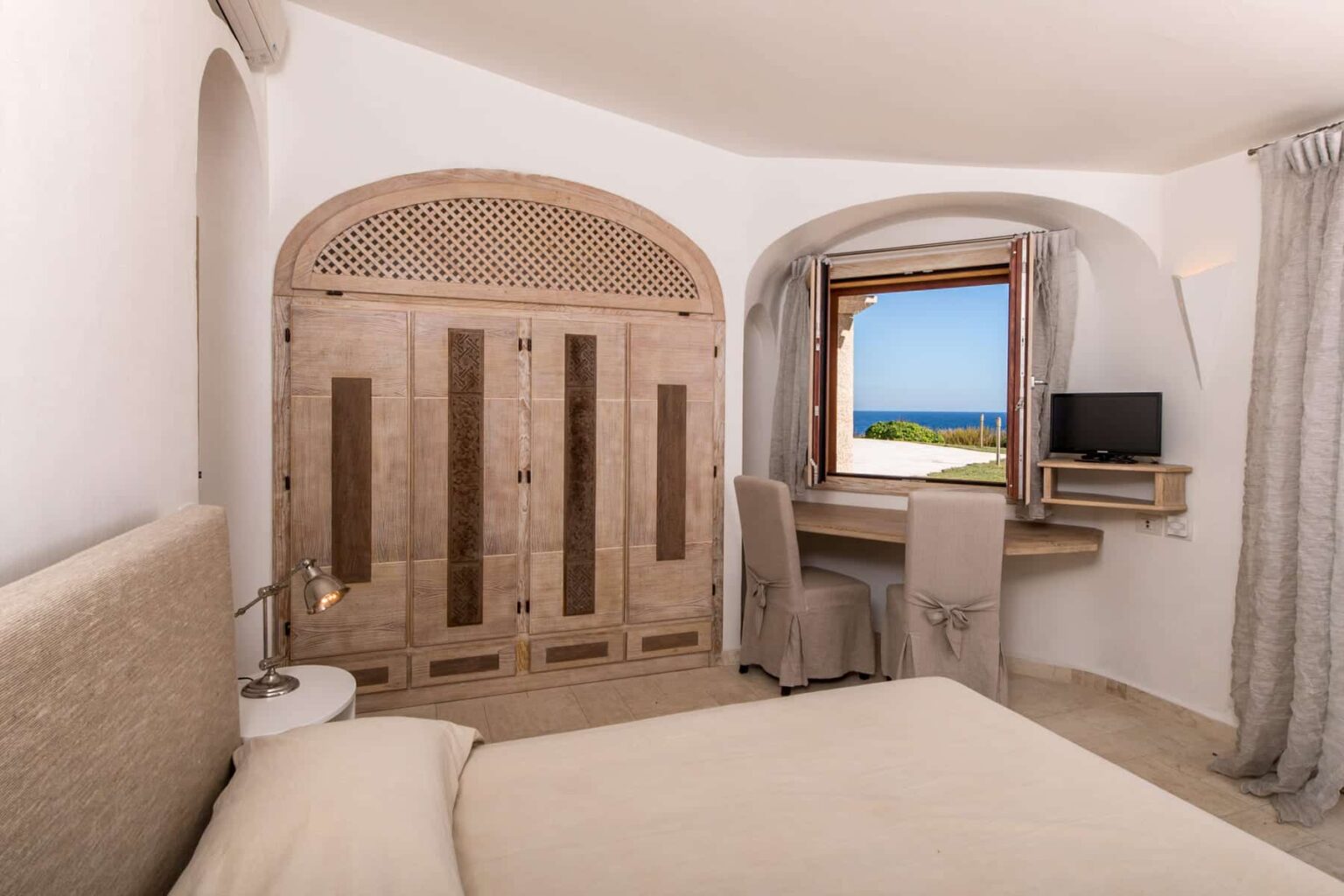 Cozy bedroom with a neutral color palette, featuring a wooden wardrobe, a desk with a small TV, and a window showcasing a scenic ocean view. Natural light floods the space, enhancing the serene atmosphere. | Only on SopranoVillas