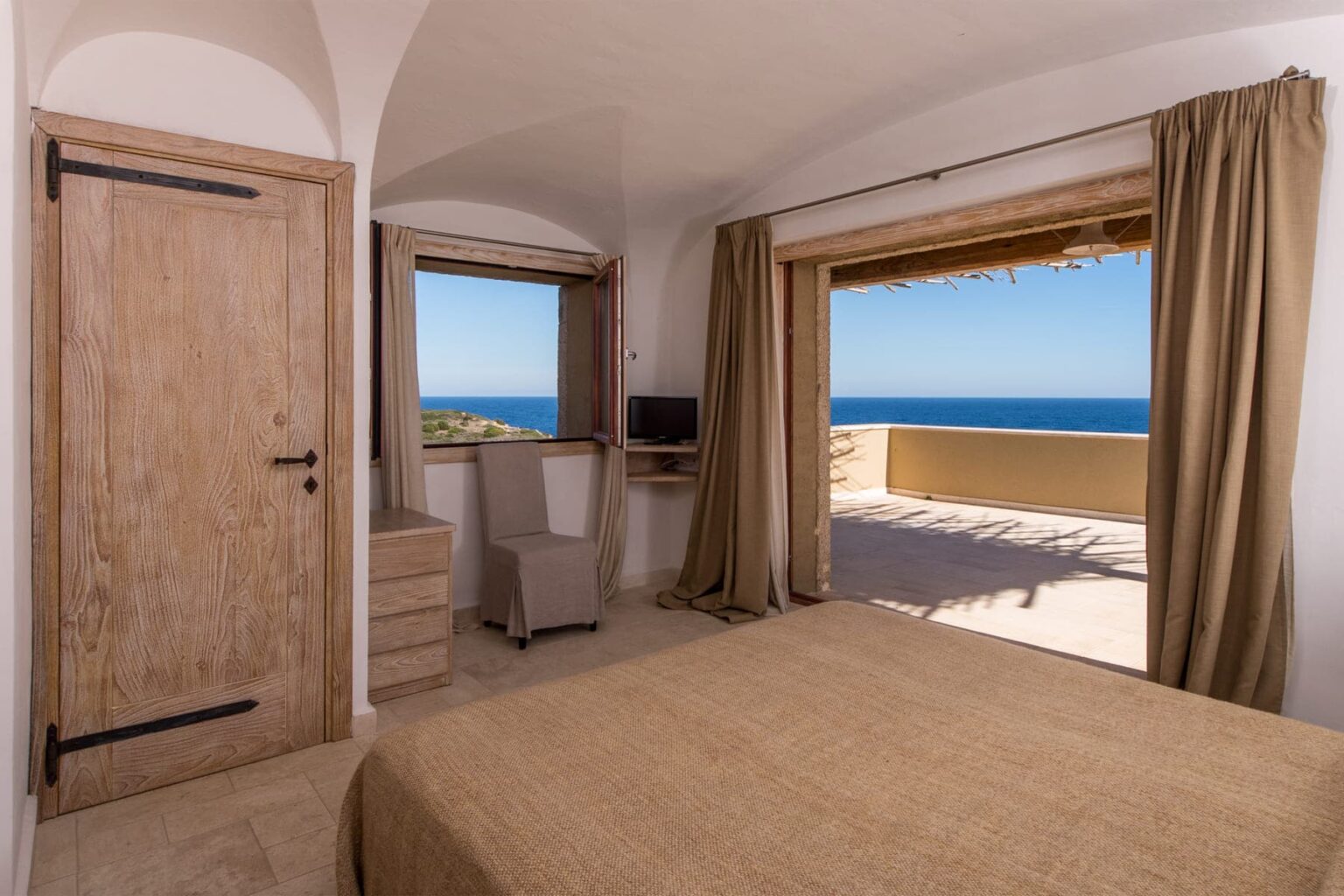 Cozy bedroom with a beige bedspread, wooden door, and a stunning ocean view through open windows. A small desk and chair are visible, creating a serene and inviting atmosphere. Ideal for relaxation and enjoying coastal scenery. | Only on SopranoVillas