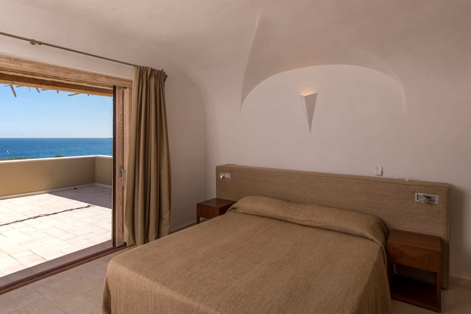 Cozy bedroom with a beige bedspread, wooden nightstands, and large windows showcasing a stunning ocean view and a private terrace. | Only on SopranoVillas