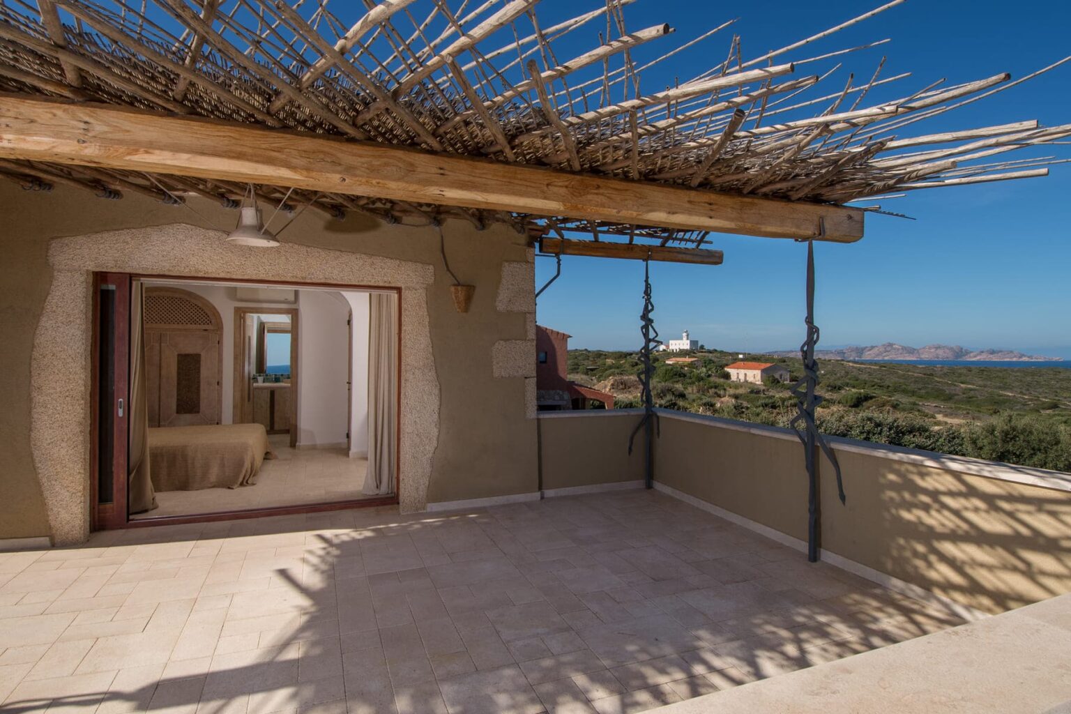 Spacious terrace with a thatched roof, offering panoramic views of the surrounding landscape and sea, leading to an interior room with a light-colored decor. Ideal for outdoor relaxation and entertaining. | Only on SopranoVillas