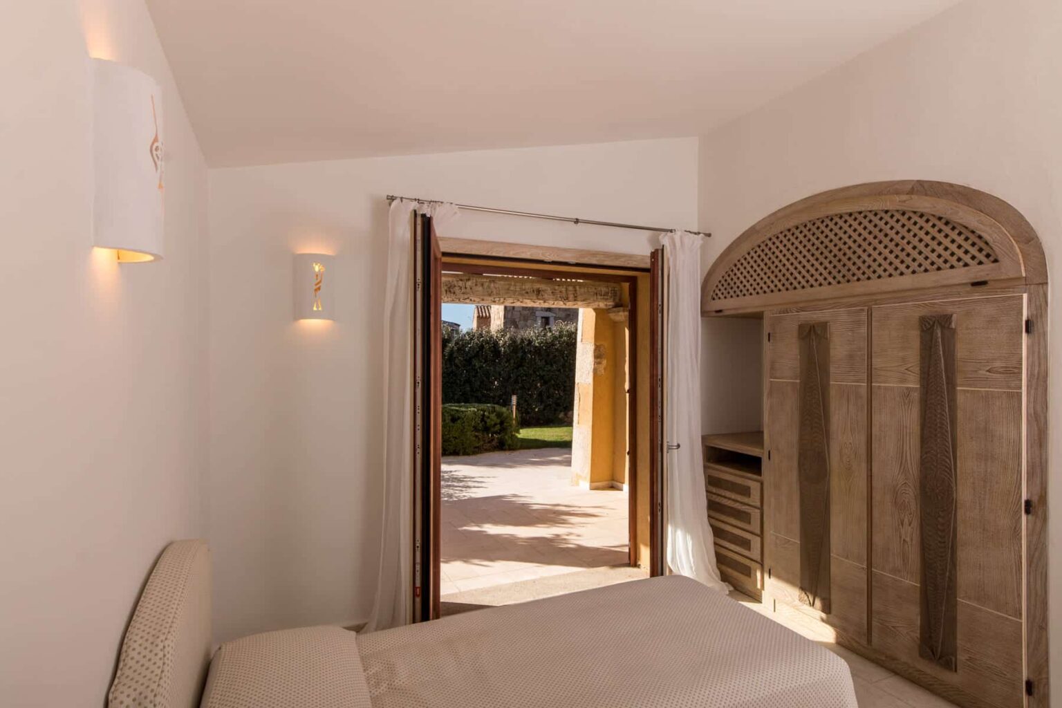 Cozy bedroom with a minimalist design, featuring a double bed, wooden wardrobe, and large sliding doors that open to a private outdoor patio surrounded by greenery. | Only on SopranoVillas
