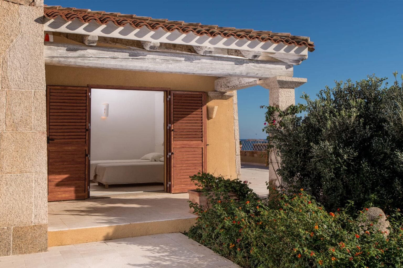 Charming exterior of a Mediterranean-style house with wooden shutters, leading to a sunlit interior and lush greenery in the foreground, under a clear blue sky. | Only on SopranoVillas