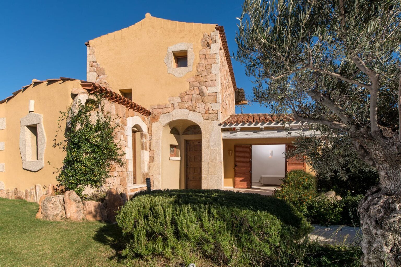 Charming rustic house with stone and stucco exterior, featuring a tiled roof, surrounded by lush greenery and shrubs, under a clear blue sky. Ideal for showcasing Mediterranean architectural style and outdoor landscaping. | Only on SopranoVillas