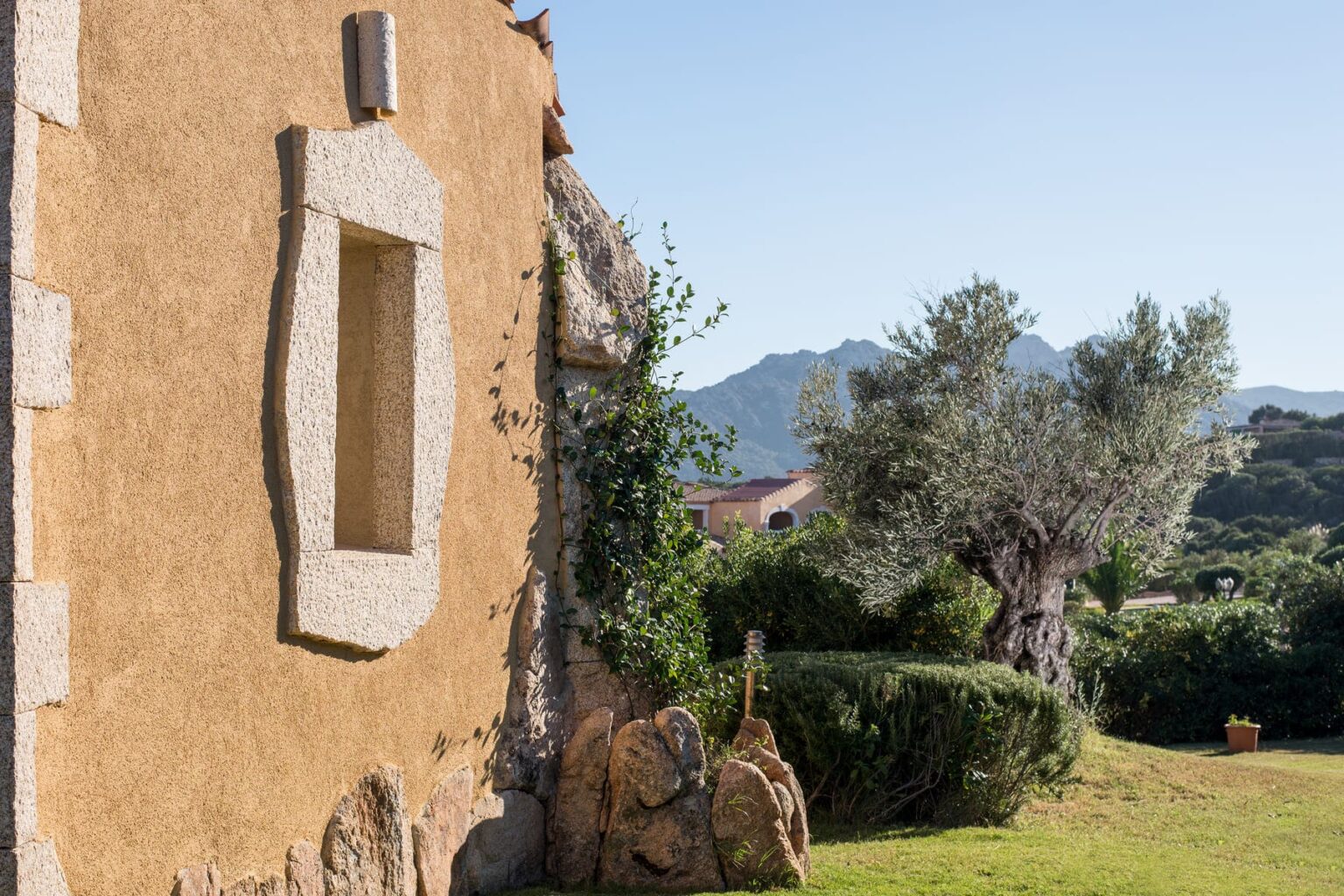 Rustic stone wall of a traditional building with a window, surrounded by lush greenery and mountains in the background, showcasing a serene landscape in a sunny setting. | Only on SopranoVillas