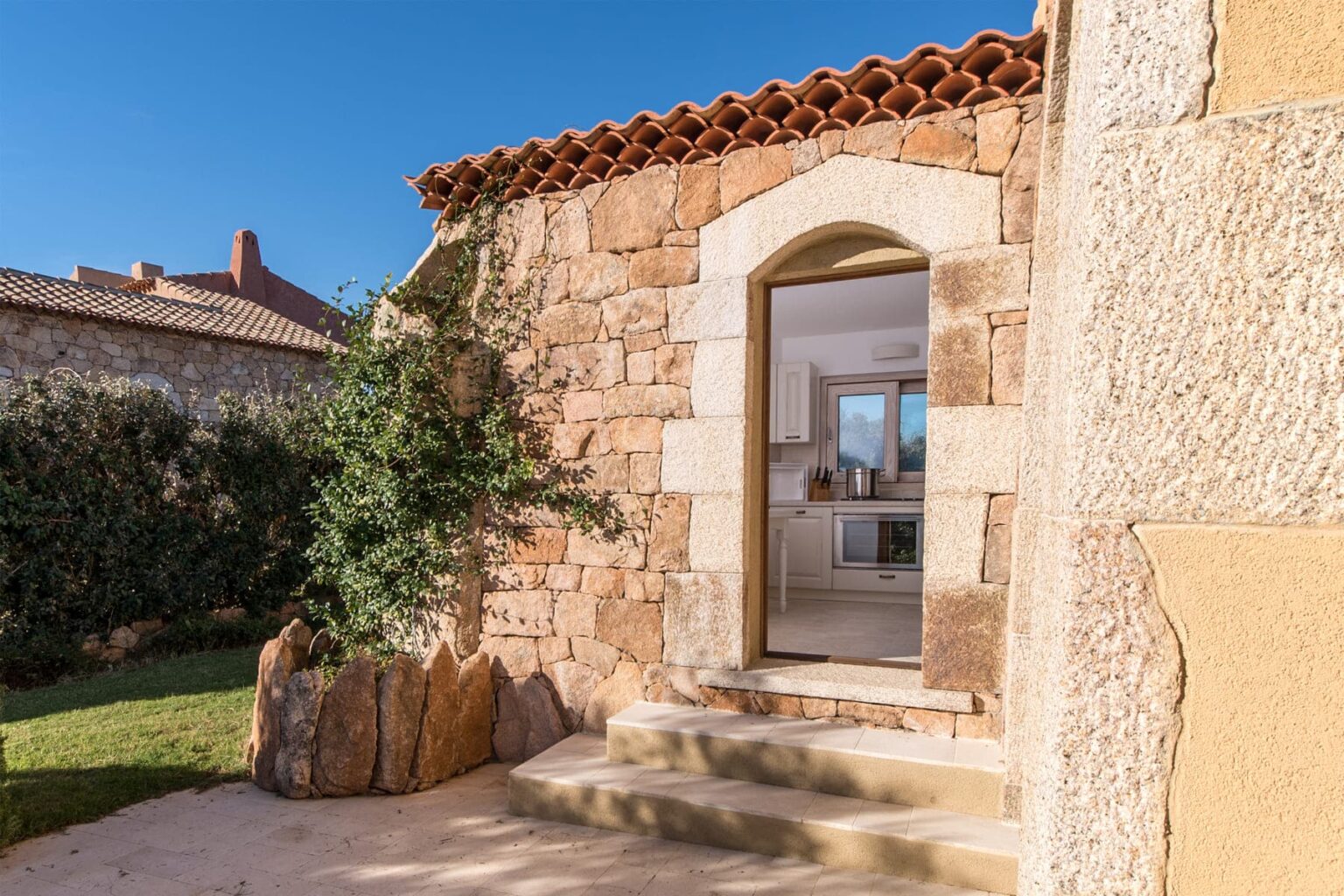 Charming stone cottage entrance with a wooden door, surrounded by lush greenery and natural stone steps, set against a clear blue sky. Ideal for showcasing rustic architecture and serene outdoor living. | Only on SopranoVillas