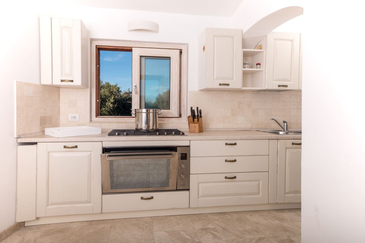 Modern kitchen interior featuring white cabinetry, a stainless steel stove, and a window with a scenic view. The countertop is equipped with a cooking pot and knife set, creating a functional cooking space. Natural light enhances the spacious feel of the kitchen. | Only on SopranoVillas