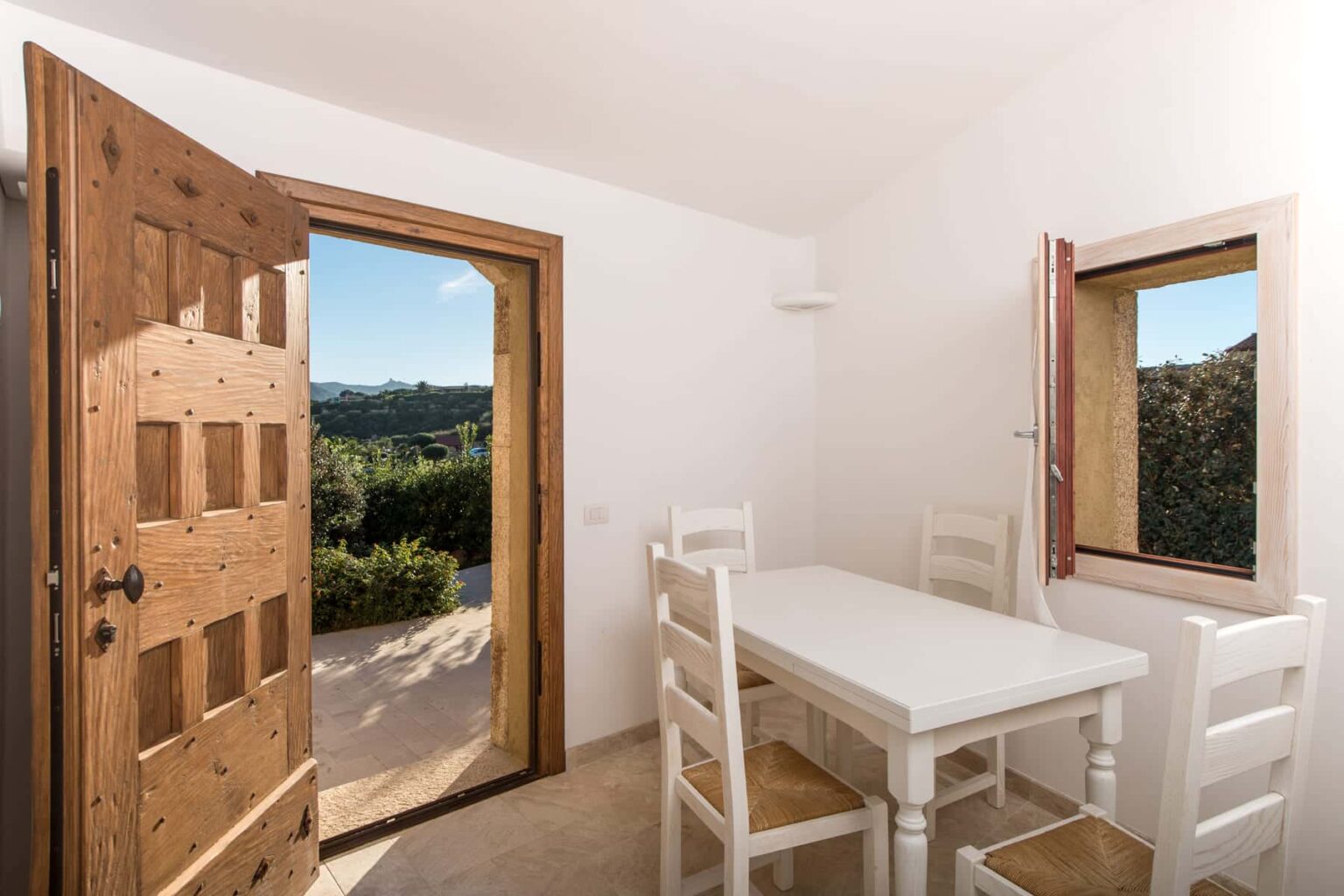 Bright and airy dining area featuring a white table and chairs, with an open wooden door leading to a scenic outdoor view. The room's minimalist design highlights natural light and creates a welcoming atmosphere. | Only on SopranoVillas