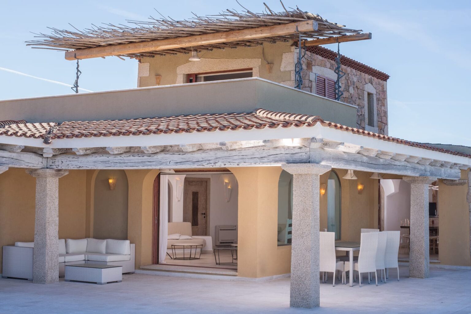 Modern Mediterranean villa with a spacious outdoor terrace, featuring elegant seating and a charming stone facade. The structure showcases a blend of traditional and contemporary architectural elements under a clear blue sky. | Only on SopranoVillas