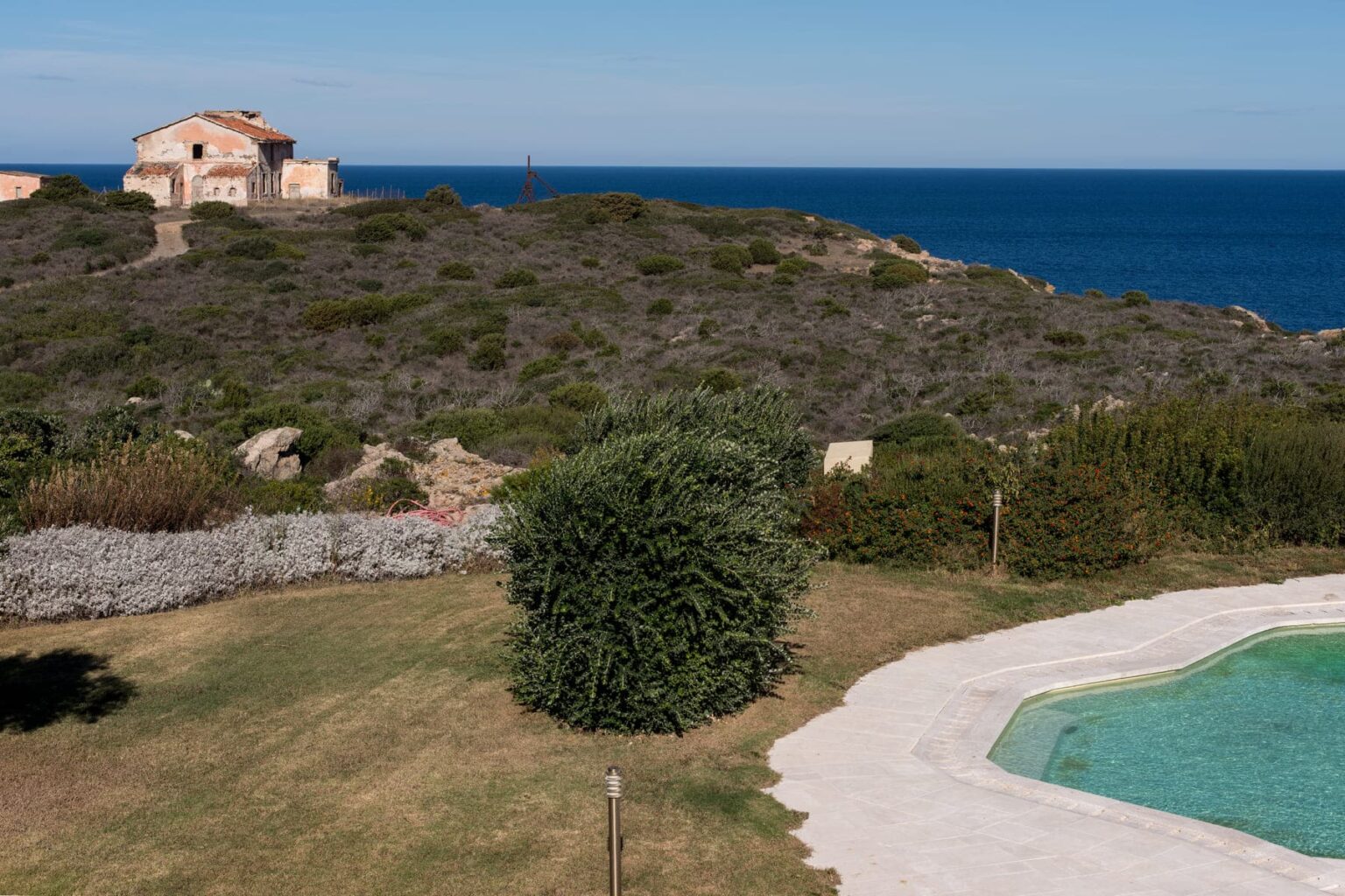 Scenic coastal view featuring an abandoned building on a hillside, lush greenery in the foreground, and a clear blue ocean in the background under a sunny sky. | Only on SopranoVillas