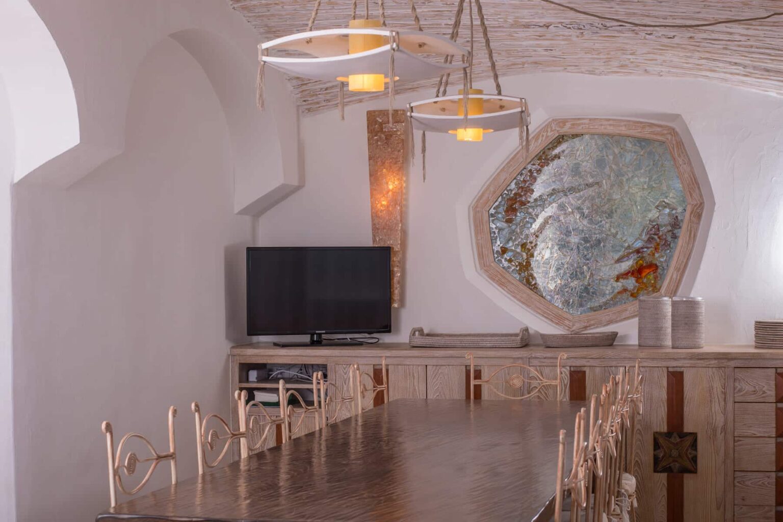 Modern dining room featuring a long wooden table with decorative chairs, a flat-screen TV, stylish pendant lighting, and an artistic octagonal mirror on the wall, set in a minimalist interior with soft white tones. | Only on SopranoVillas