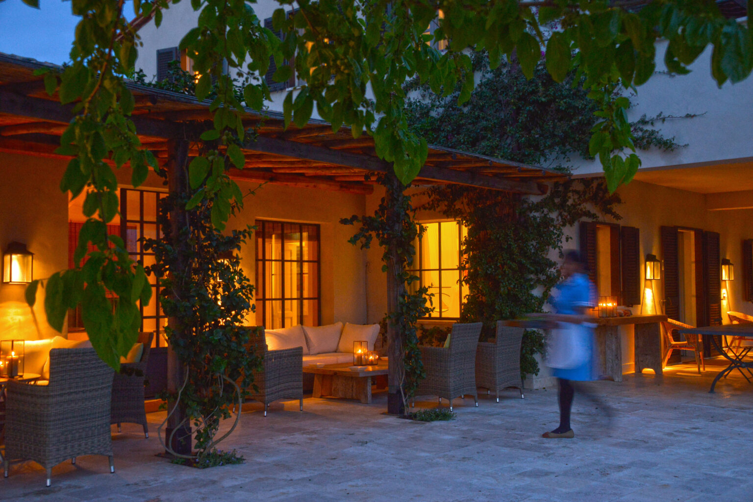 Cozy outdoor seating area of a boutique hotel, featuring wicker chairs and warm lighting, with a staff member serving guests in the evening. Lush greenery and glowing windows enhance the inviting atmosphere. | Only on SopranoVillas
