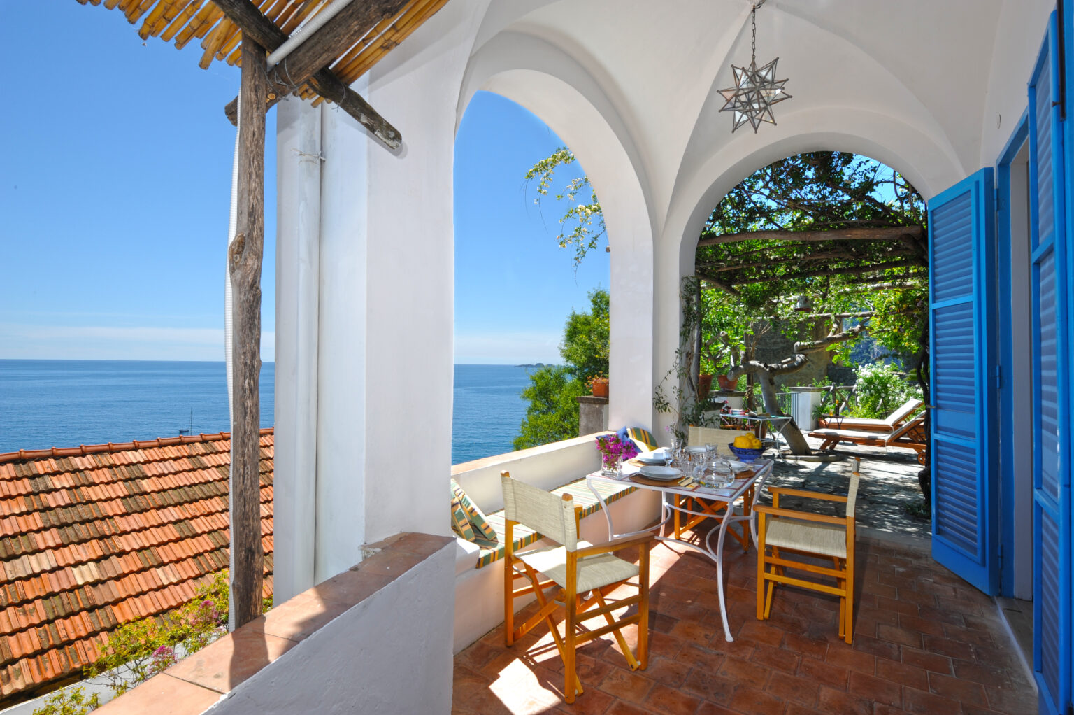 Cozy outdoor dining area with a stunning view of the ocean, featuring a table set for a meal, comfortable chairs, and vibrant greenery. The space is adorned with a decorative light fixture and surrounded by arched walls and blue shutters, creating a serene atmosphere perfect for relaxation. | Only on SopranoVillas