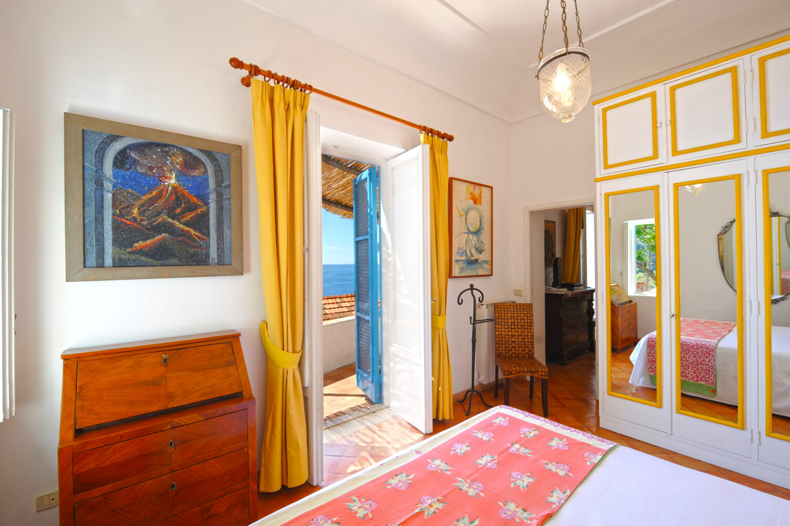Bright and airy bedroom featuring a wooden dresser, yellow curtains, and a blue-framed window with a sea view. The room includes a colorful artwork of a volcano, a patterned bedspread, and a mirror reflecting the inviting decor. | Only on SopranoVillas