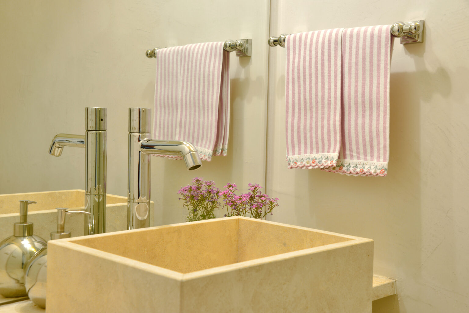 Modern bathroom sink with polished chrome faucets, striped pink towels hanging on towel racks, and small purple flowers in a decorative pot, creating a stylish and inviting ambiance. | Only on SopranoVillas