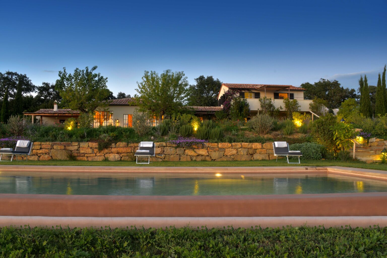 Serene view of a modern villa surrounded by lush greenery, featuring a swimming pool in the foreground, cozy lounge chairs, and warm outdoor lighting against a twilight sky. Ideal for luxury vacation rentals or serene getaways. | Only on SopranoVillas