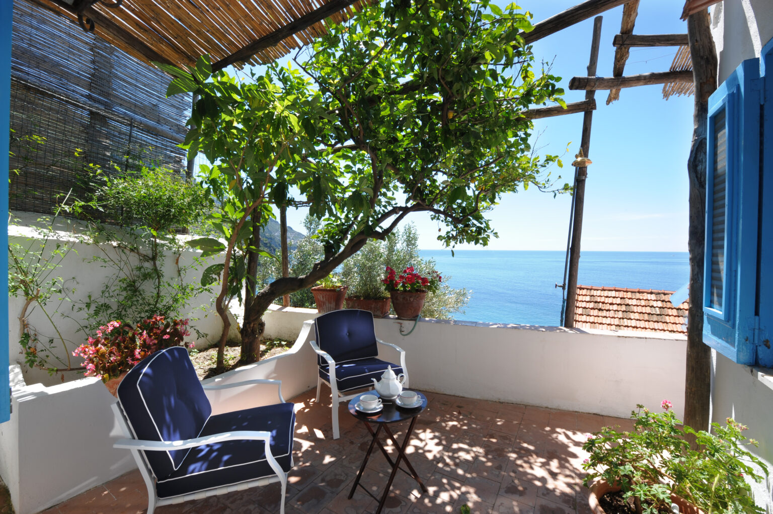 Cozy outdoor terrace with blue chairs and a small table set for tea, surrounded by greenery and overlooking the ocean. Ideal spot for relaxation and enjoying scenic views. | Only on SopranoVillas