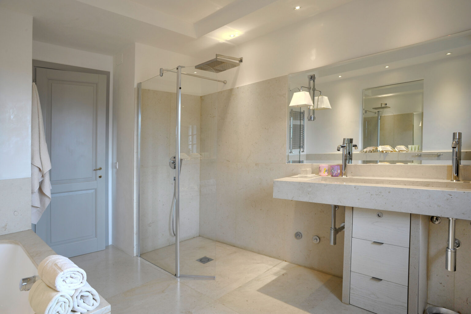 Modern bathroom featuring a glass shower, dual sinks with sleek chrome faucets, and a minimalist design. The space includes a bathtub, neatly rolled towels, and soft lighting, creating a serene and stylish atmosphere. | Only on SopranoVillas