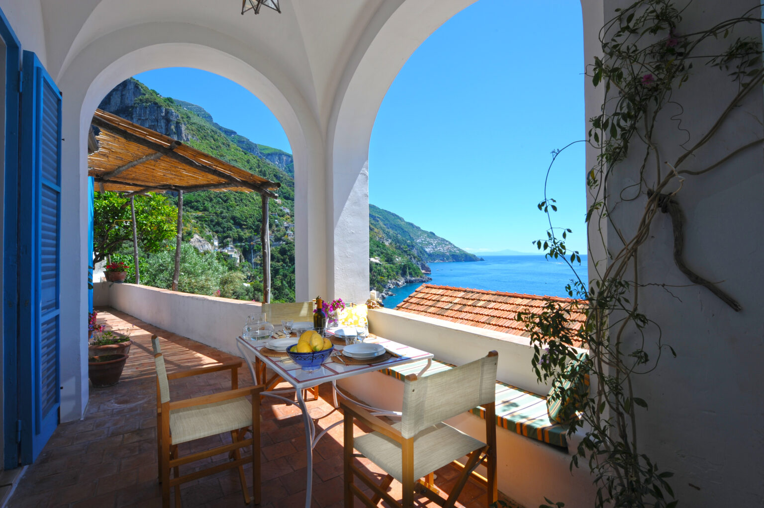 Scenic view from a terrace overlooking the Amalfi Coast, featuring a dining table set with dishes, lemons, and flowers, framed by arches and lush greenery. | Only on SopranoVillas